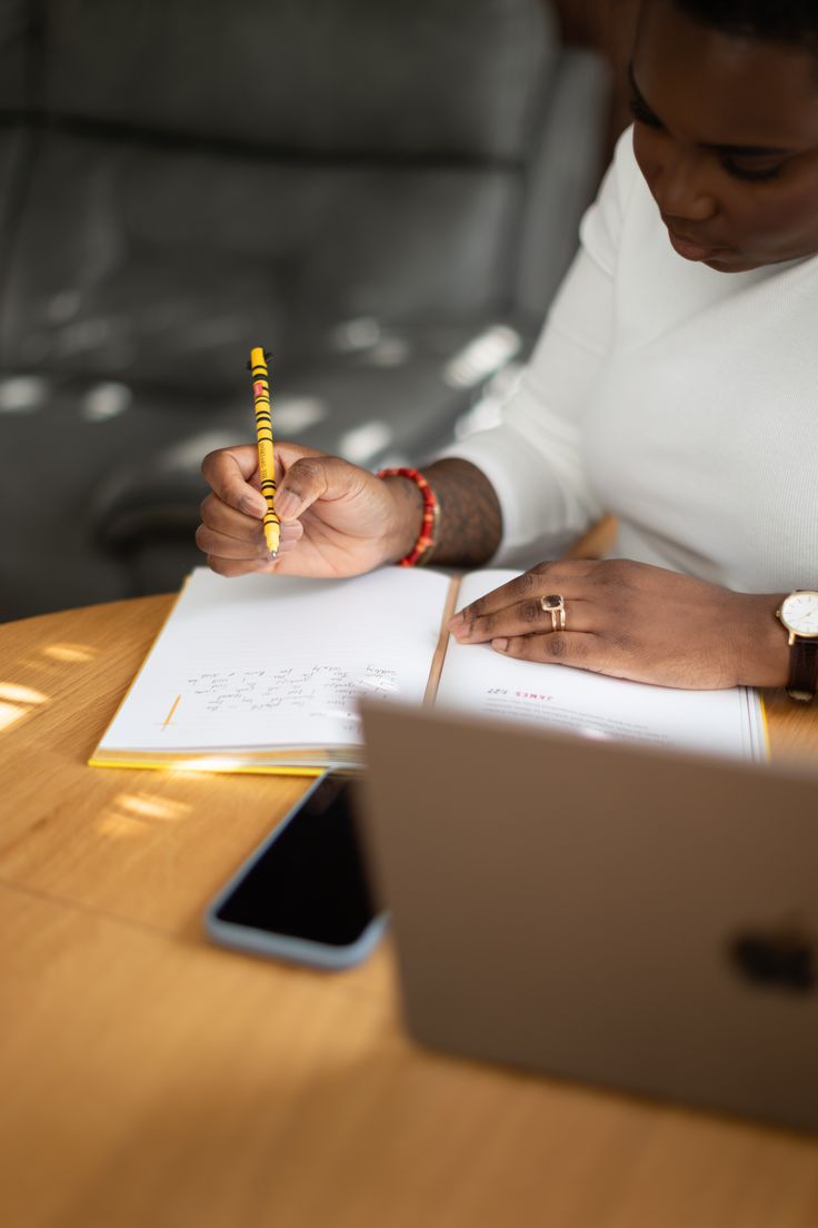 Pessoa escrevendo em um caderno em uma mesa com laptop e celular